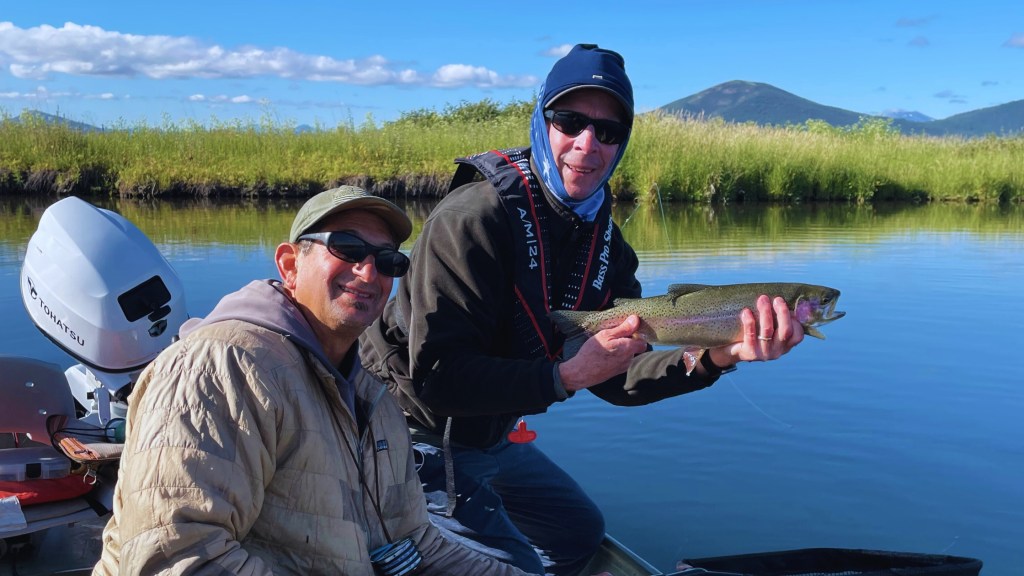 Two smiling anglers pose on a calm river in Northern California, one proudly holding a freshly caught rainbow trout, with grassy banks and distant mountains under a clear blue sky